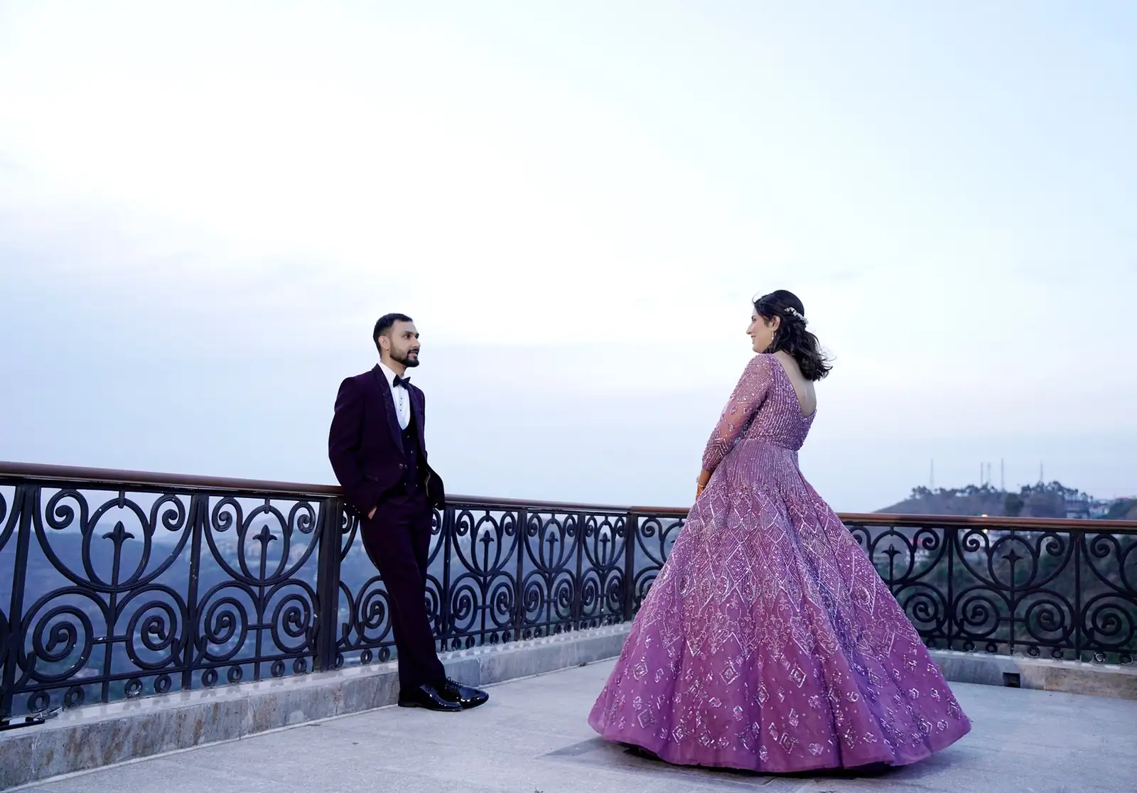 Elegant couple in formal wear on a balcony, bride in purple gown, groom in suit. 