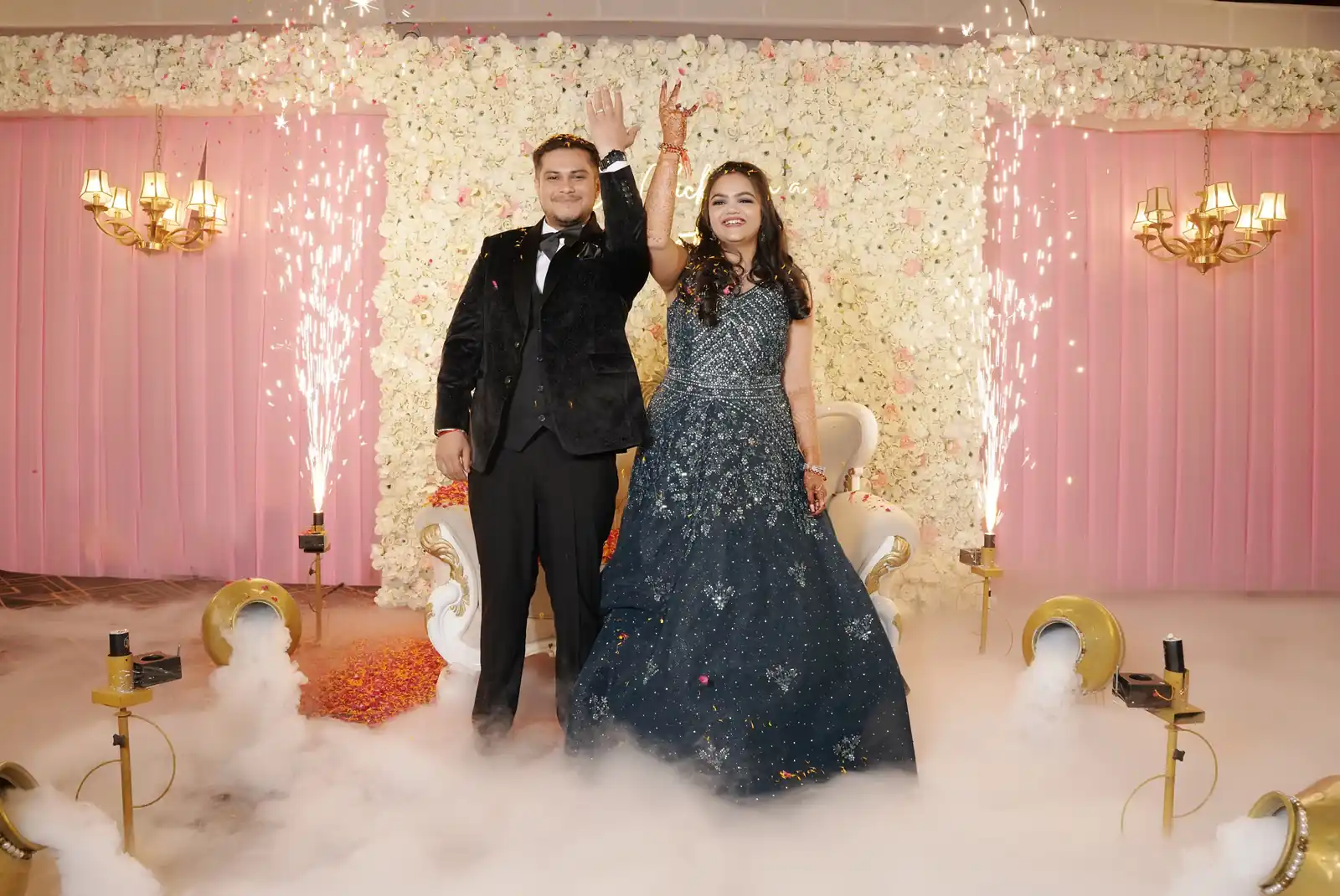 Couple celebrating engagement with fireworks and fog machine on stage with floral backdrop.
