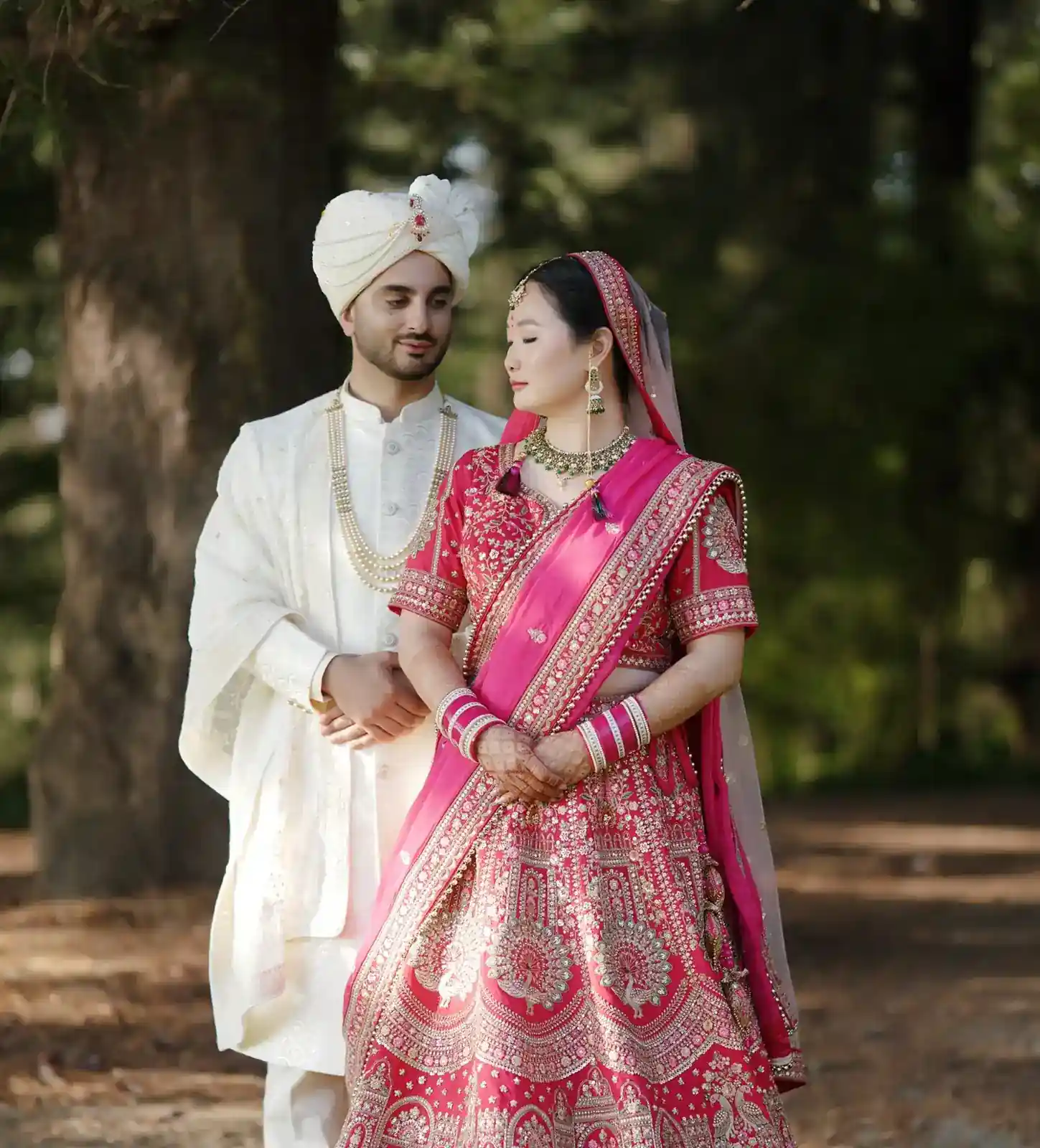 Indian wedding: Bride in red lehenga and groom in white sherwani embrace in outdoor setting.