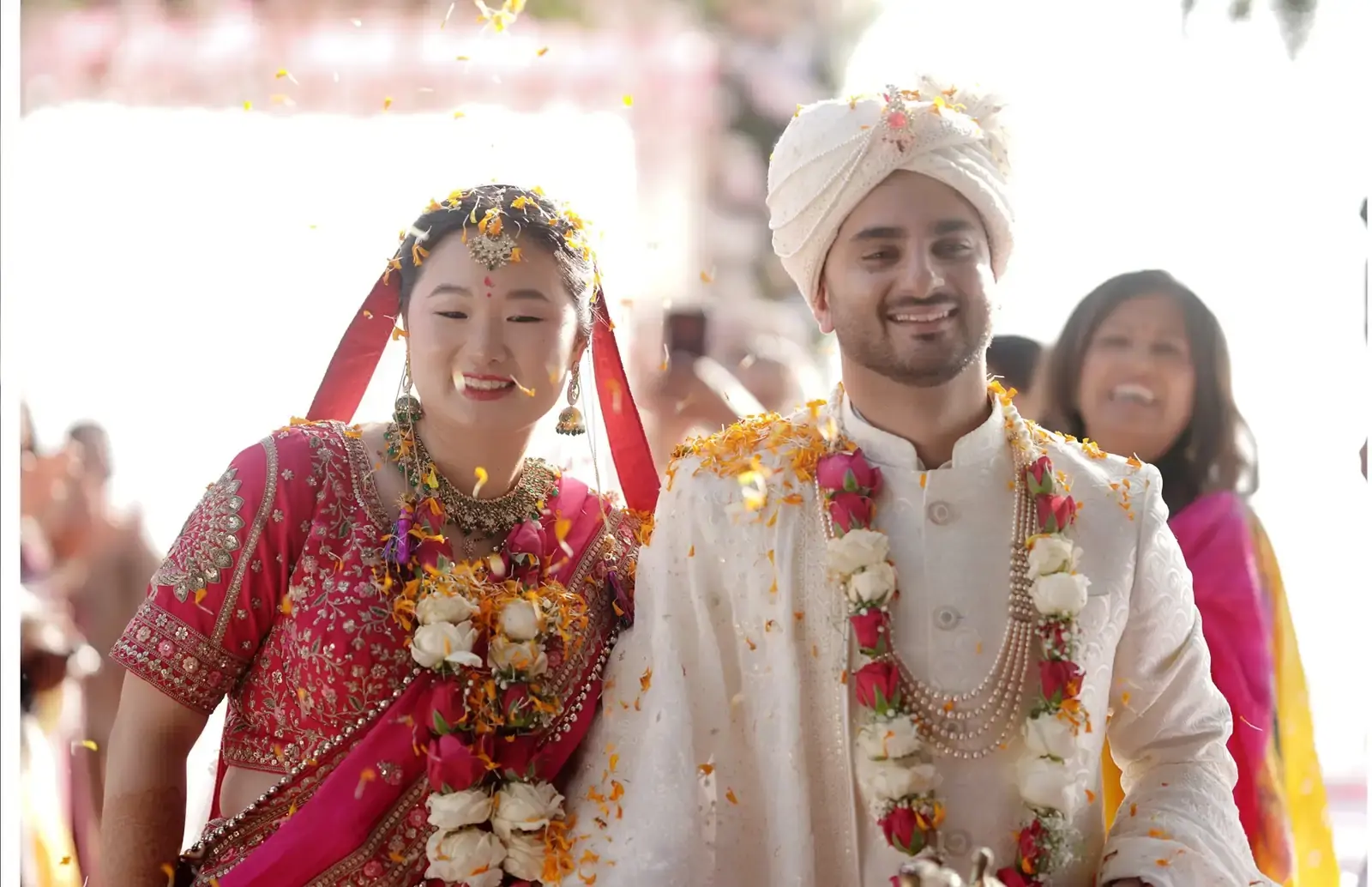 Happy couple at Indian wedding, bride in red sari, groom in white sherwani, flower petals falling.
